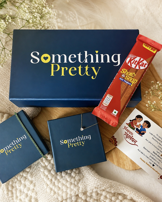 A curated gift set from Something Pretty, featuring a navy blue gift box, a matching notebook, a small jewelry box with a necklace, a KitKat snack bar, and a card, arranged on a wooden board with white flowers over a cream knit blanket.
